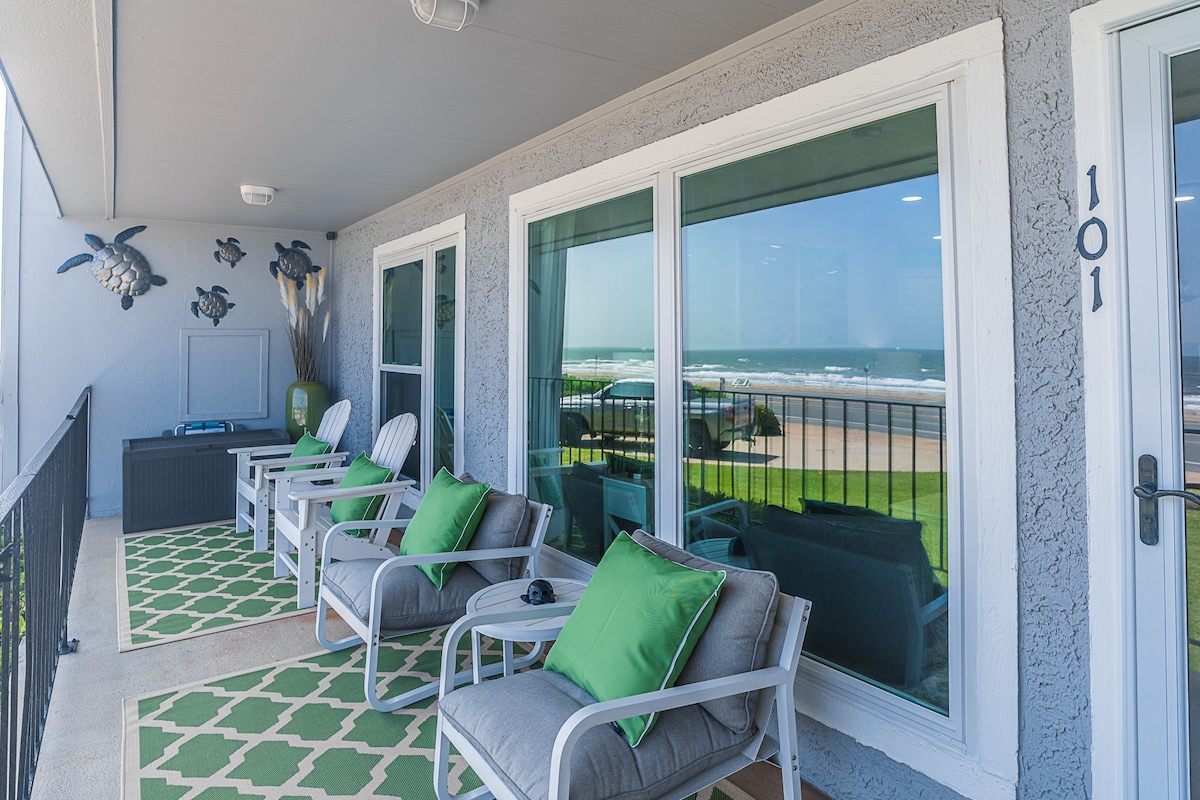 A spacious balcony features comfortable seating, including several lounge chairs and small tables. Green cushions add a pop of color, while ocean views are visible through large windows, highlighting the proximity to the beach.