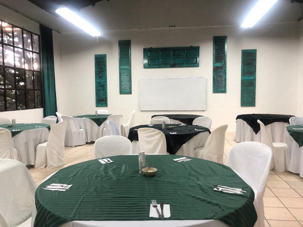 A spacious dining area features round tables set with green tablecloths, complemented by white chairs. Bright overhead lights illuminate the room, highlighting the textured walls adorned with decorative green shutters. A whiteboard stands at one end, offering a versatile space for gatherings.