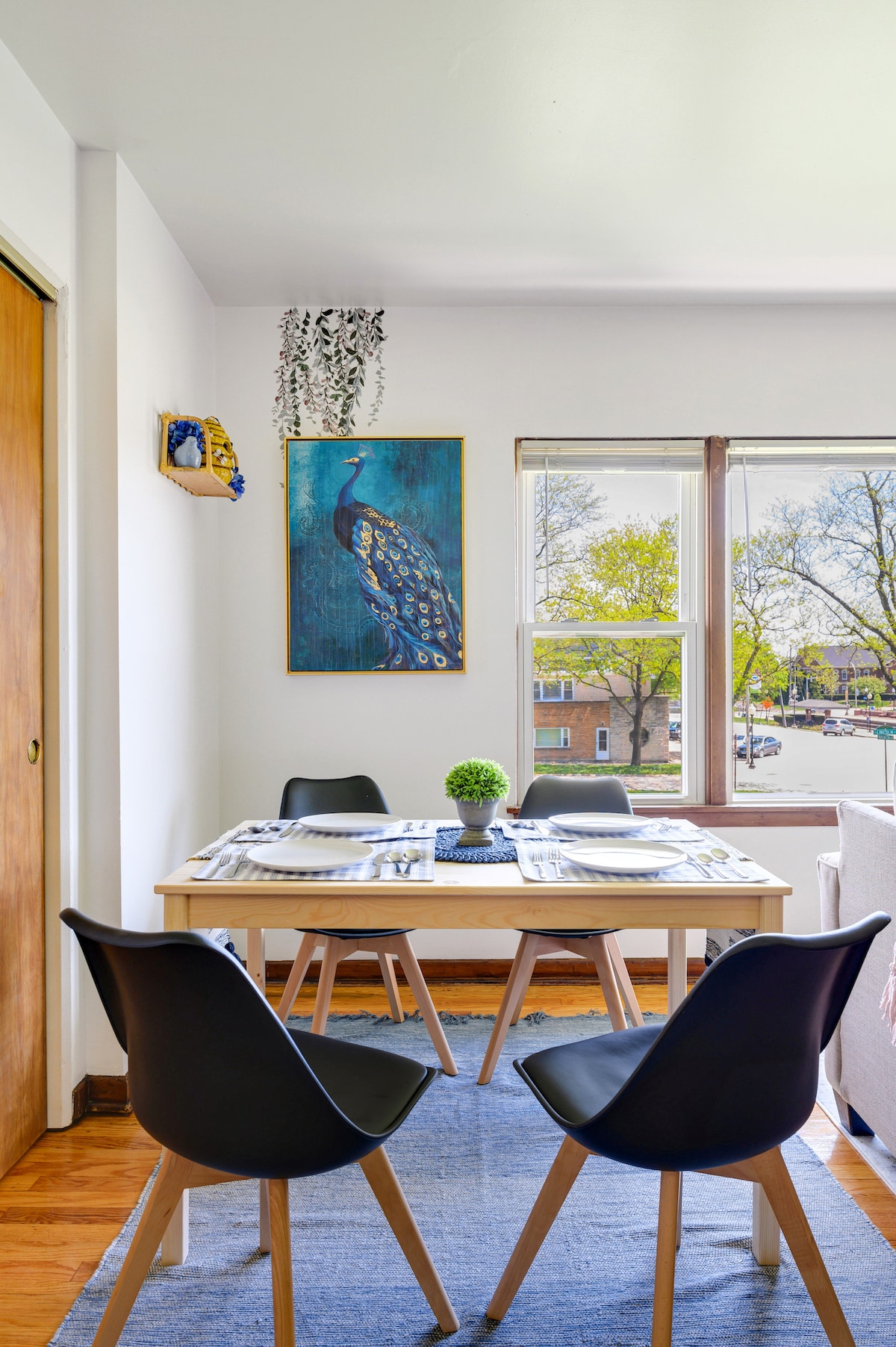 A dining area features a light wood table set for four, surrounded by sleek black chairs. A vibrant peacock artwork adorns the wall above the table, while a small potted plant adds a touch of greenery. Natural light filters through large windows, illuminating the space.