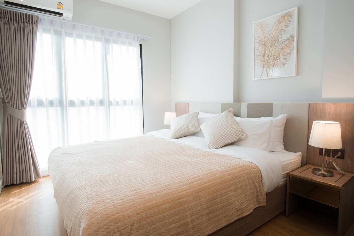 A cozy bedroom features a spacious bed with light bedding and decorative pillows. Natural light filters through sheer curtains covering a large window. A bedside table with a lamp is positioned on either side of the bed, complementing the minimalistic decor.