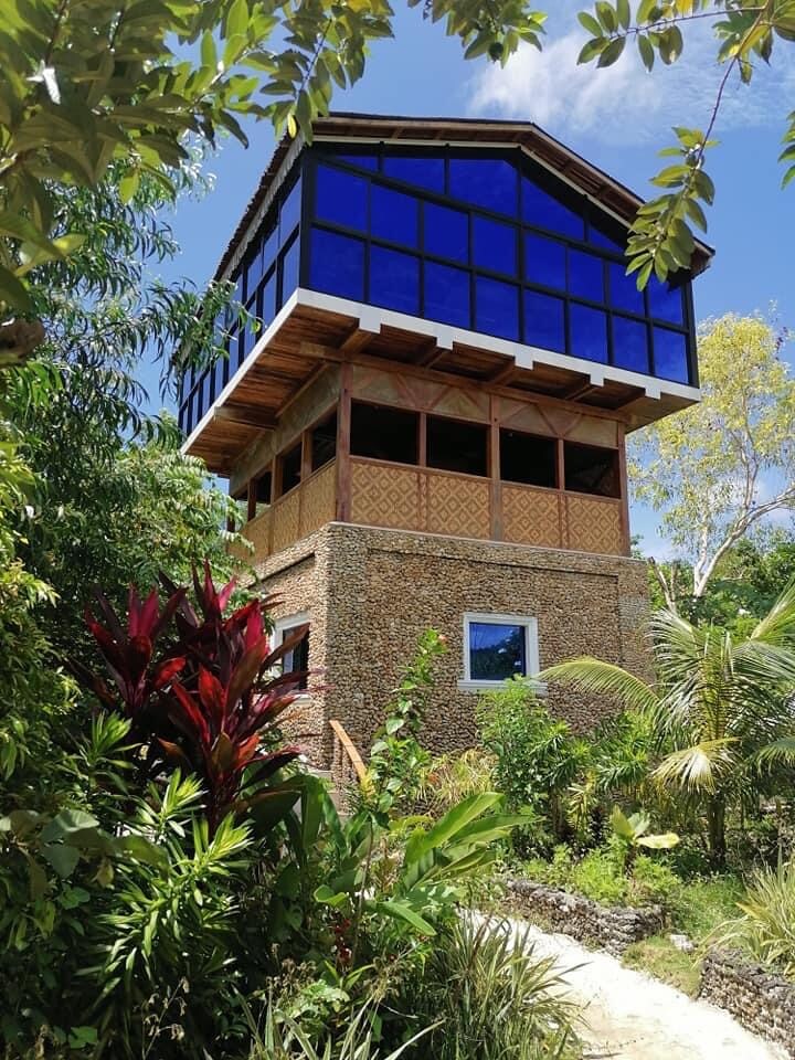 A multi-level structure is surrounded by lush greenery, featuring large blue glass windows that allow natural light to fill the interior. The lower portion presents a textured stone facade, while colorful tropical plants frame the pathway leading up to the building.