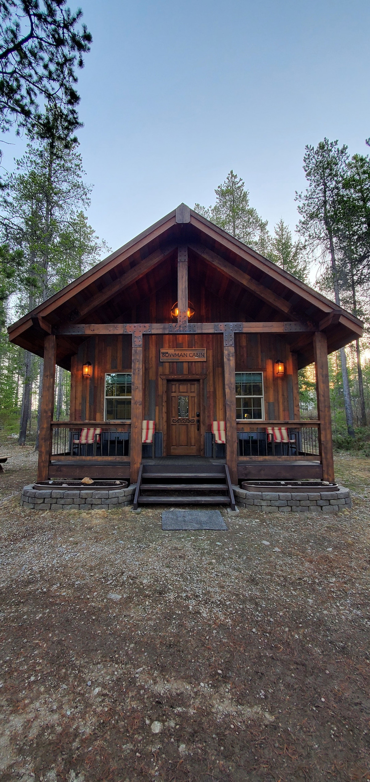 The rustic cabin features a large front porch with comfortable seating on both sides. The entrance displays a sturdy wooden door framed by warm lighting. Surrounding trees create a natural setting, adding to the cabin’s peaceful ambiance.