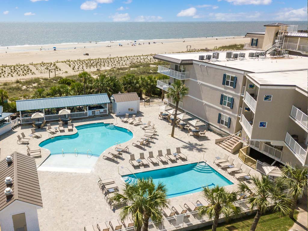 Aerial view of the resort complex showcasing two swimming pools surrounded by lounge chairs. The beach and ocean are visible in the distance, with rows of palm trees lining the pool area, offering a relaxing atmosphere. The bright sky complements the inviting outdoor space.