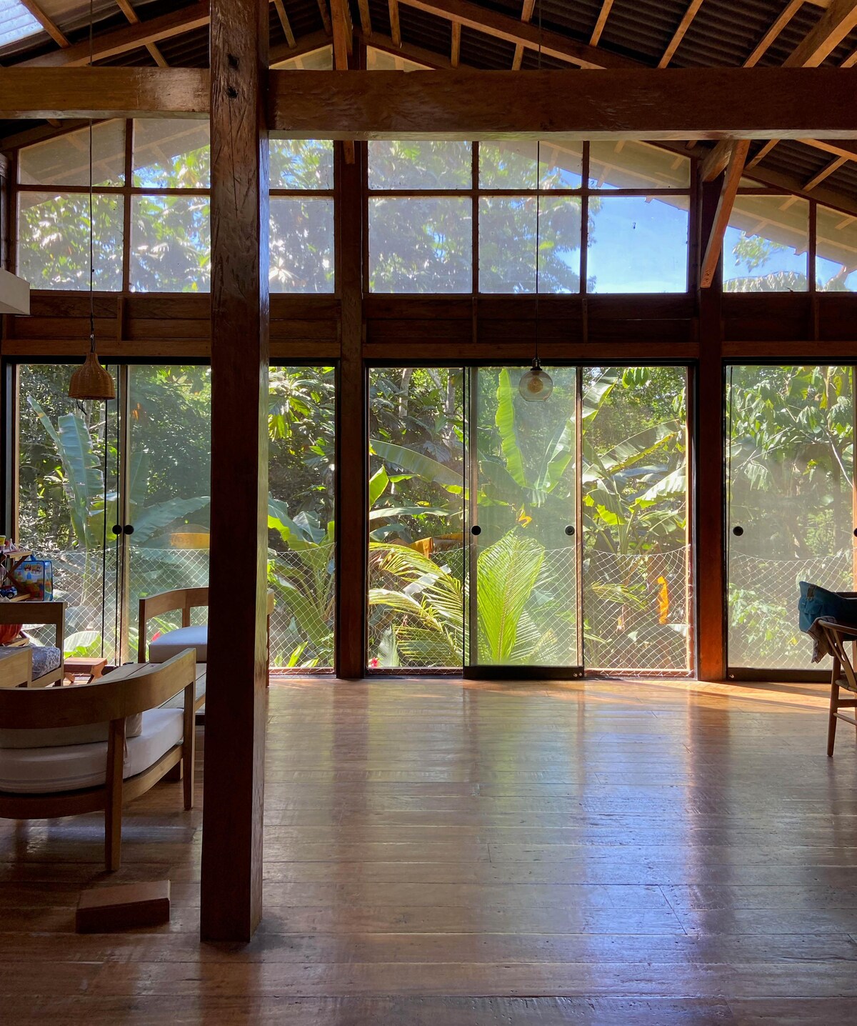 A spacious living area is illuminated by natural light through large glass doors, offering views of lush tropical greenery. Wooden beams and floors contribute to the rustic charm. Nearby furniture includes a cozy seating arrangement. The atmosphere reflects a connection with the surrounding nature.