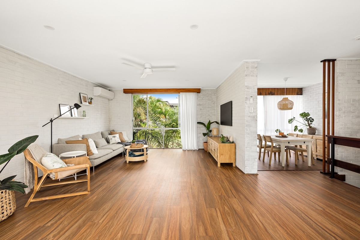 A spacious living area features light-colored walls and warm wooden flooring. A comfortable seating arrangement is highlighted by a modern armchair and a relaxed sofa. Nearby, a dining area is visible, featuring a wooden table surrounded by chairs, with large windows offering natural light.