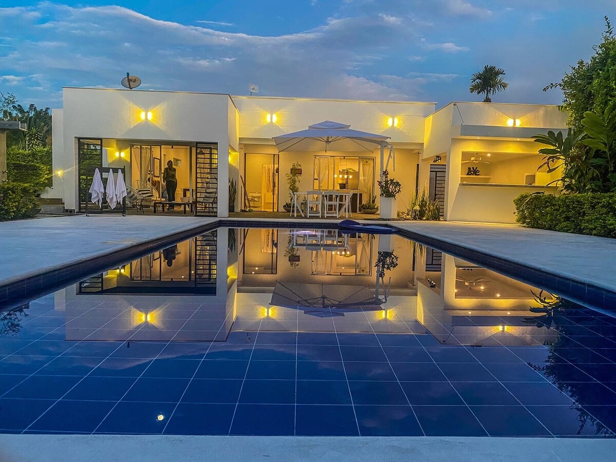 A modern structure with large glass doors opens to an outdoor space featuring a tranquil pool. The pool's deep blue tiles reflect the soft glow of evening lights from the building. An umbrella and dining area create an inviting atmosphere for relaxation and gatherings.