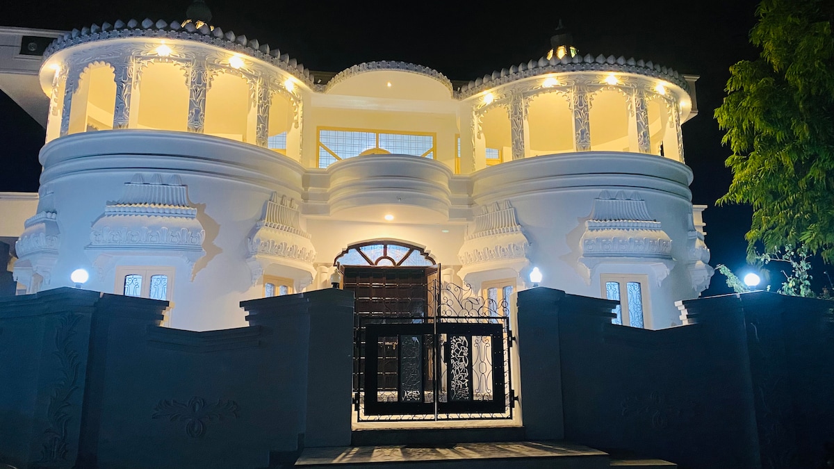The elegant exterior of the homestay is highlighted by ornate architectural details and soft lighting, creating a welcoming atmosphere. The entrance features a gated entryway surrounded by landscaped greenery, complemented by decorative stonework and a grand façade that reflects a blend of traditional and modern styles.
