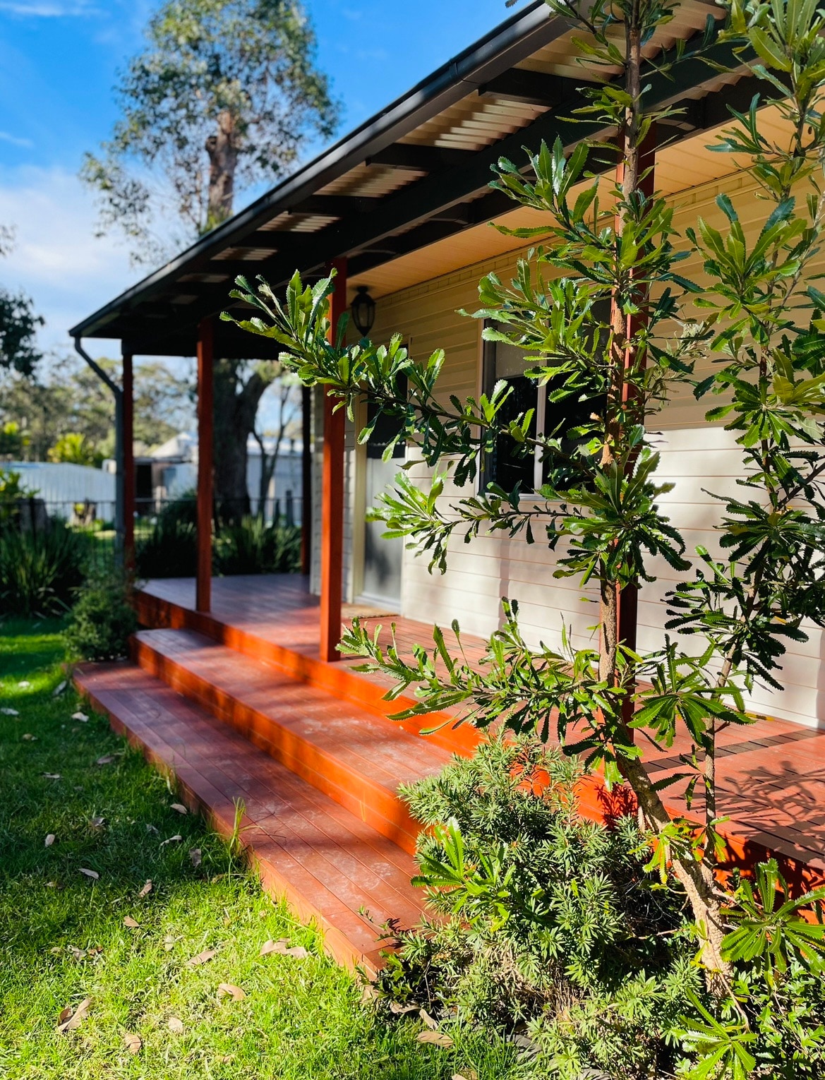 The exterior of the cottage is highlighted by a wooden deck and steps leading up to the entrance. Lush greenery frames the space, promoting a serene environment. A clear sky is visible above, suggesting a bright and inviting atmosphere.