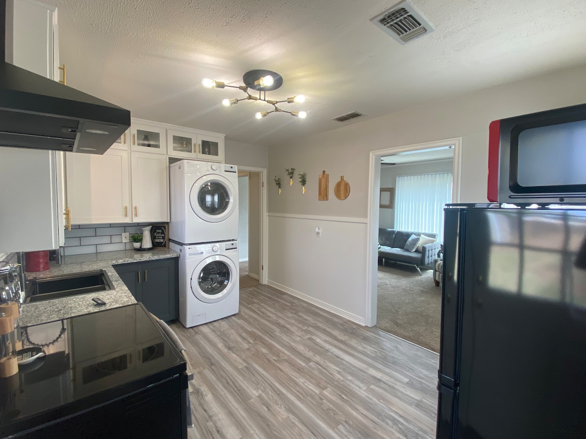A modern laundry area is featured, showcasing stacked washing and drying machines. A black refrigerator is also present. Light fixtures offer illumination over gray laminate flooring. A doorway leads to a separate living space, where natural light filters through sheer curtains.