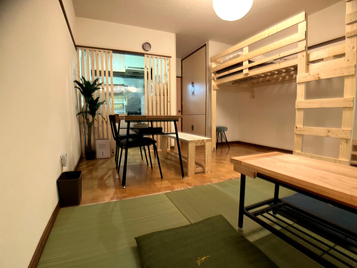 A living area features a wooden table and chairs set beside a low coffee table. A lofted bed is visible in the background, with light wood finishes. A green tatami mat covers the floor, and a potted plant adds a touch of greenery to the space.