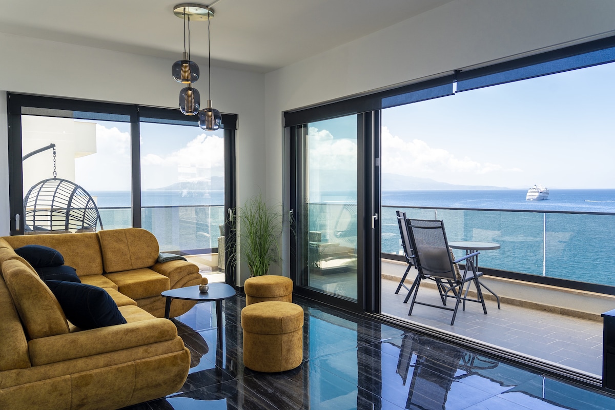 A modern living space is highlighted by large glass doors offering expansive sea views. A plush, mustard-colored sofa with matching ottomans provides comfortable seating. A small round table sits in the center, while outdoor seating and a balcony are noted beyond the glass doors.