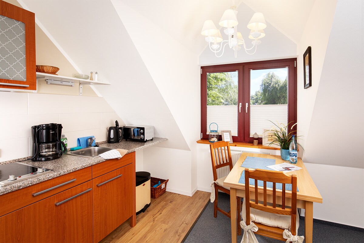 A bright kitchen area features wooden cabinetry and a granite countertop with modern appliances. A dining table is positioned near a large window, allowing natural light to fill the space. A chandelier hangs from the ceiling, adding a warm touch to the room.
