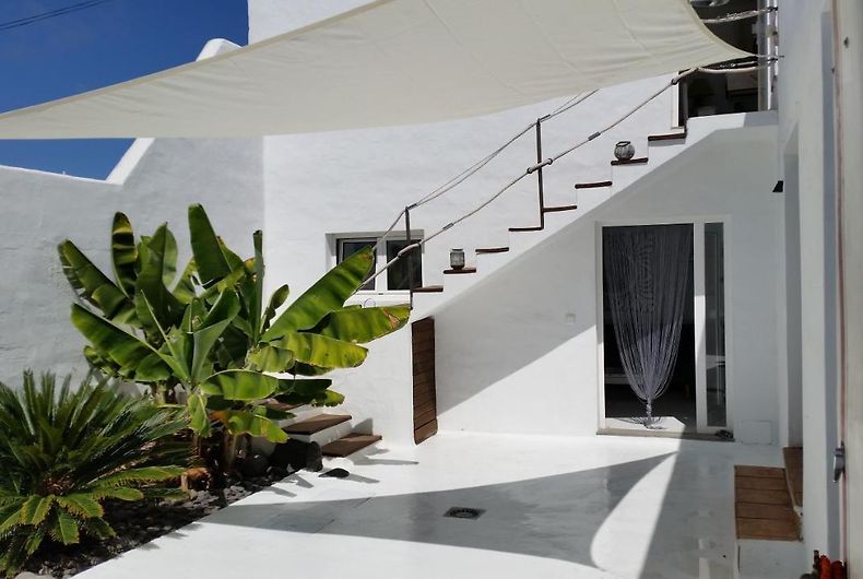 An outdoor patio area features a blend of greenery and clean white walls. Sturdy stairs lead to the upper level, topped with a shade sail. A glass door provides access to the interior, while potted plants add subtle touches of nature.
