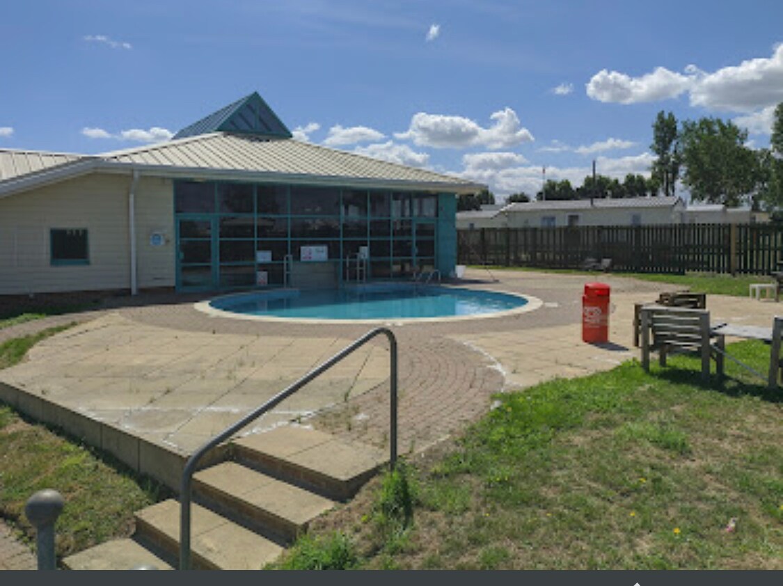 A clear blue swimming pool is surrounded by a spacious patio area, featuring steps leading down to the water. Adjacent to the pool, a large building with glass windows offers indoor facilities. Green grass and a wooden fence frame the background.