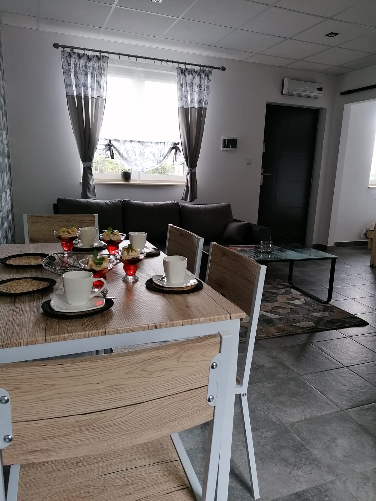A modern living space features a dining table set for four, complete with cups and decorative fruit. A comfortable sofa is positioned nearby, with light filtering through the windows accented by patterned curtains. The room is designed with neutral tones and sleek tiles.