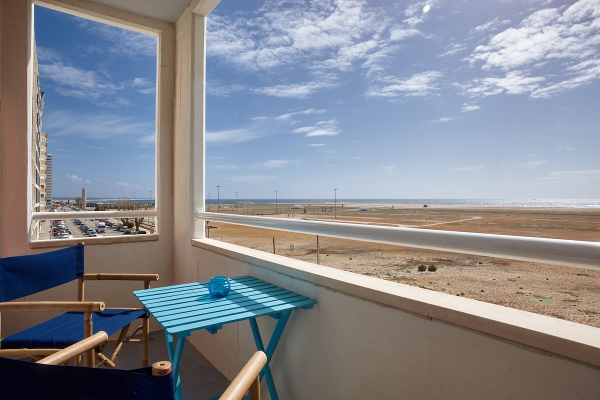 A private balcony offers a clear view of the beach and ocean under a bright blue sky, with sunlight illuminating a turquoise table. Two wooden chairs provide a comfortable seating area, inviting relaxation while enjoying the coastal scenery.