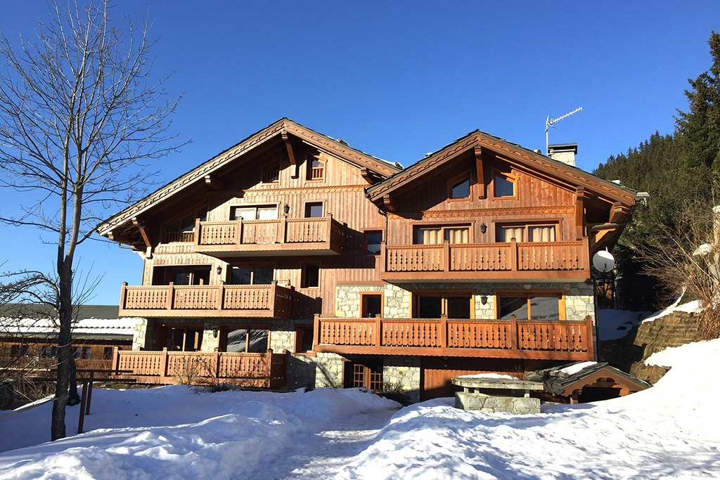 A charming alpine building is set against a clear blue sky, showcasing wooden architecture with multiple balconies. Snow blankets the ground, while trees and a mountain backdrop enhance the natural setting. The inviting exterior reflects traditional chalet style.