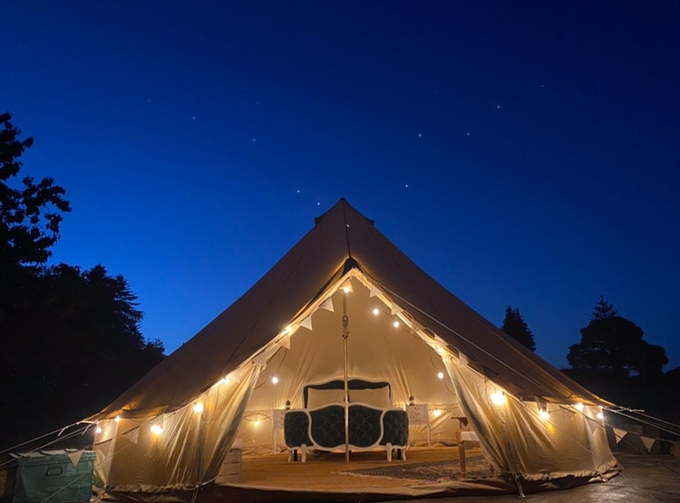 A spacious bell tent is illuminated by soft outdoor lights, standing under a night sky filled with stars. A comfortable sofa chair is visible inside, creating a cozy retreat. The tent's shape is accentuated by the clear backdrop of evening twilight.