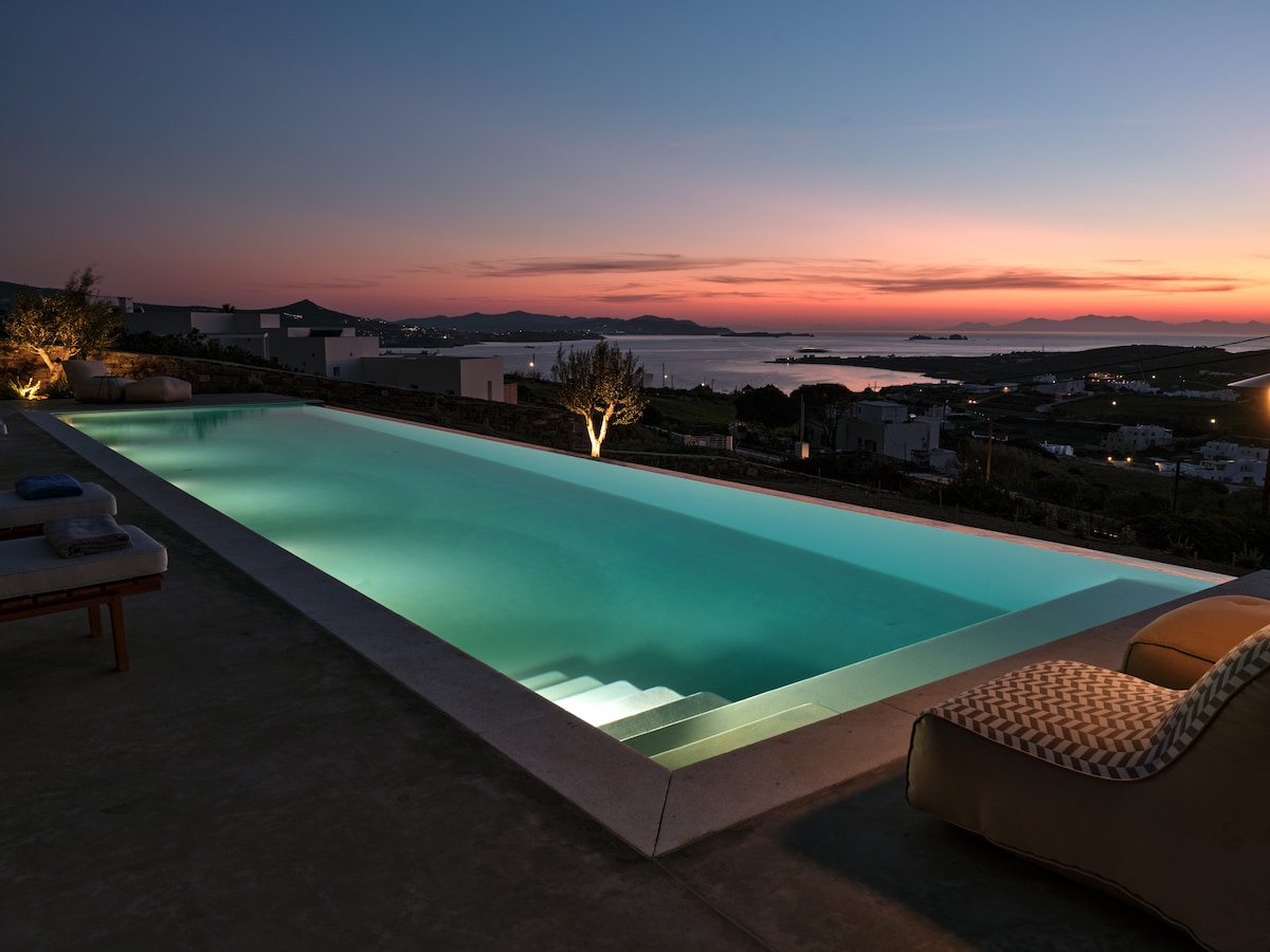 A serene infinity pool is illuminated by soft lighting, extending towards a scenic view of the hills and sea at sunset. The calming turquoise water reflects the evening sky, while steps lead gently into the pool. Nearby lounge chairs offer a comfortable place to relax.