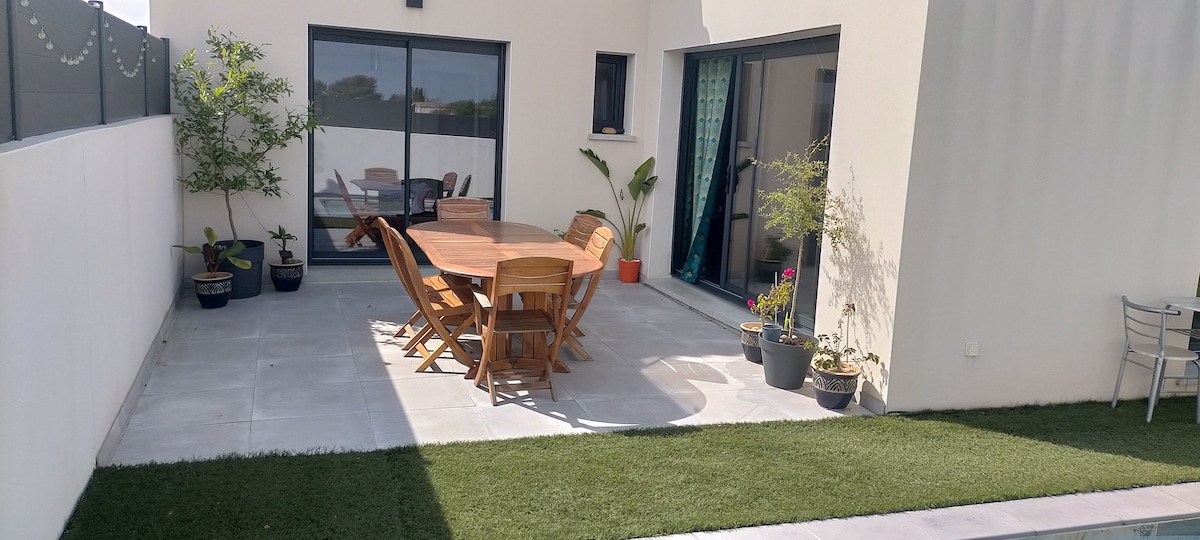 An outdoor patio area features a wooden dining table surrounded by six chairs. Potted plants line the space, complementing the lawn area. Large glass doors provide access to the interior, while a canopy of bright curtains adds a touch of color.