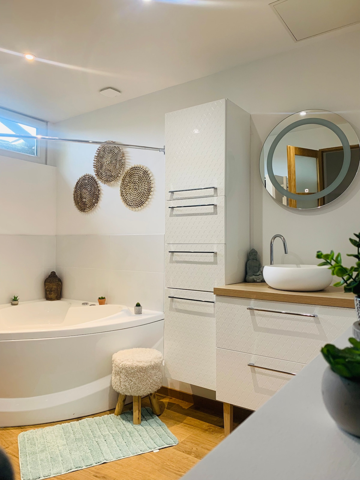 The bathroom features a modern design with a rounded bathtub and a stylish round mirror above a sink. A wooden stool and decorative wall elements add interest. Natural light filters through a window, highlighting the neutral colors and minimalist decor.