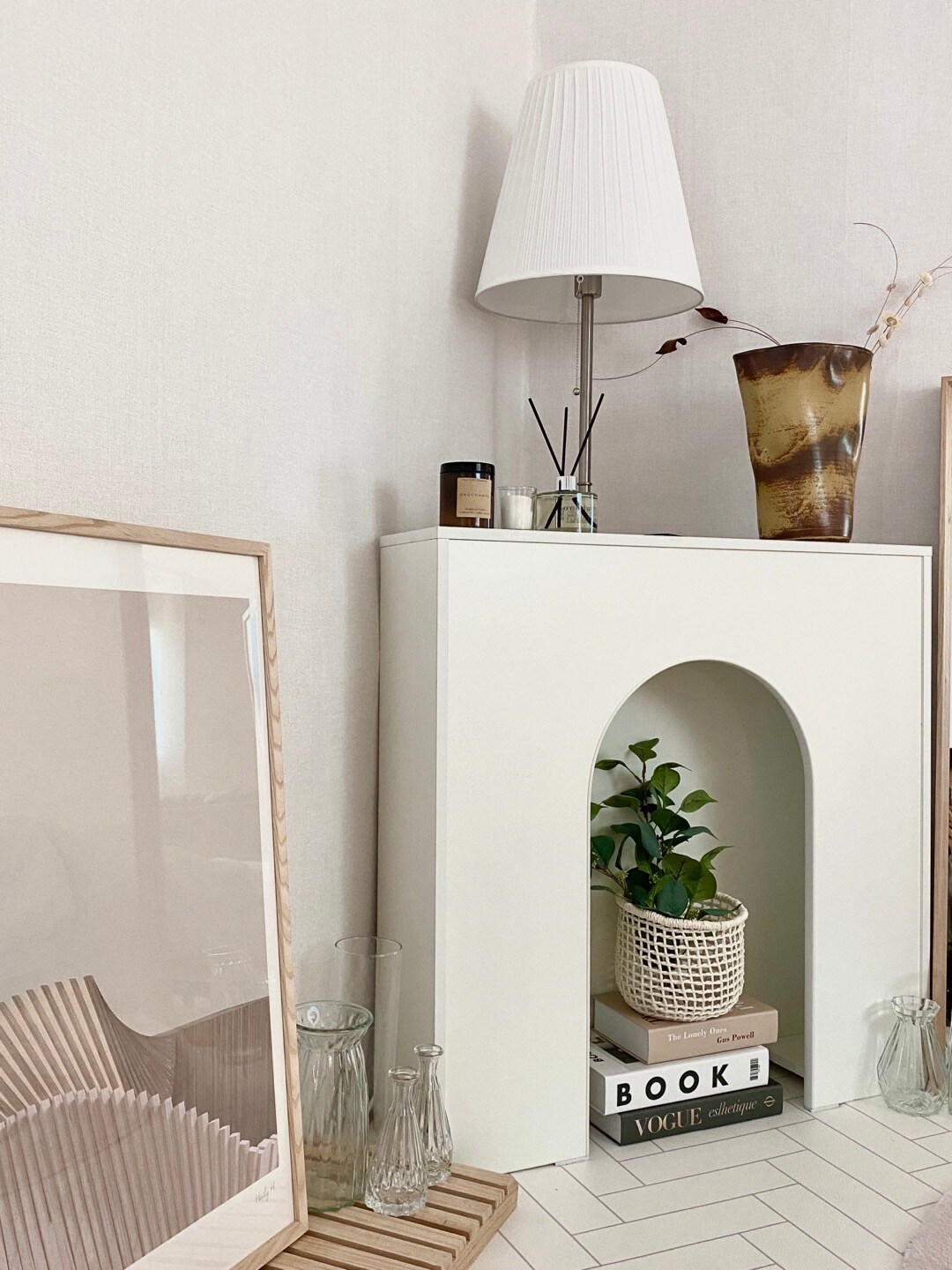 A minimalistic corner features a white shelf with an arch design. A small plant is housed in a woven basket. A modern lamp with a pleated shade stands beside it, accompanied by decorative vases and stacked books. A framed artwork leans against the wall.