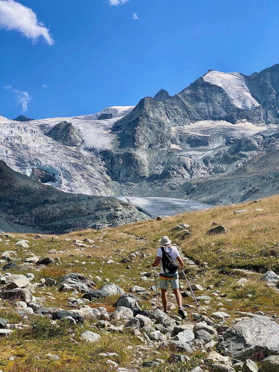 A hiker is seen navigating through a rocky landscape, surrounded by expansive mountain peaks and a glacier in the distance. The clear blue sky enhances the natural scenery, with patches of grass and scattered boulders covering the ground.