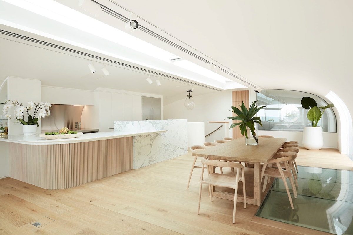 A modern open-plan kitchen and dining area is highlighted by natural wood flooring. A large dining table with wooden chairs is complemented by a marble-topped kitchen island. Potted plants add freshness, while ample natural light floods through skylights and glass doorways.