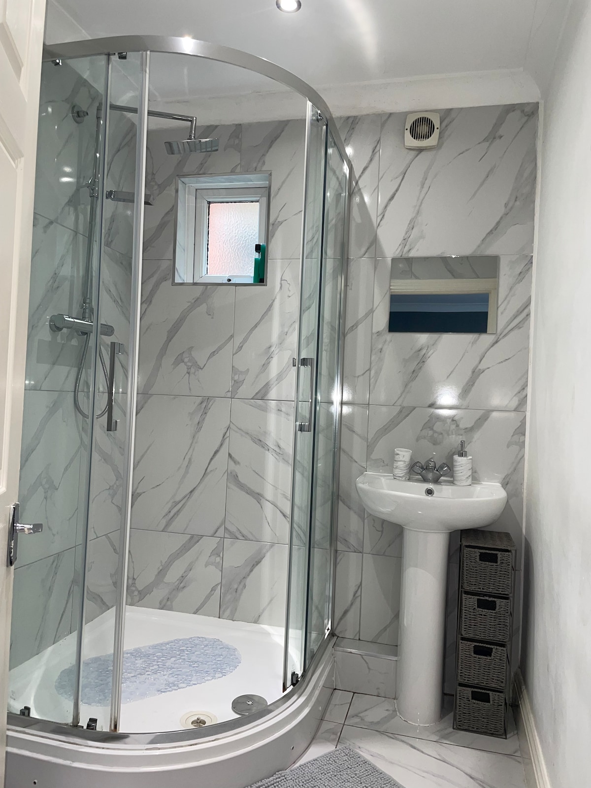 A bathroom features modern styling, with a curved glass shower enclosure set against marble-patterned tiles. A compact pedestal sink is positioned nearby, complemented by a wall mirror. A light blue mat rests on the floor, adding a touch of color to the neutral space.