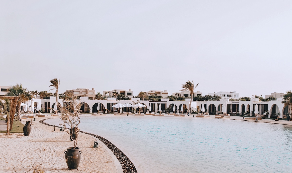 A serene pool area encircled by elegant arches and palm trees is visible. Comfortable loungers are placed along the sandy beach, and white umbrellas provide shade. The tranquil water reflects the calm sky, creating a peaceful atmosphere for relaxation.