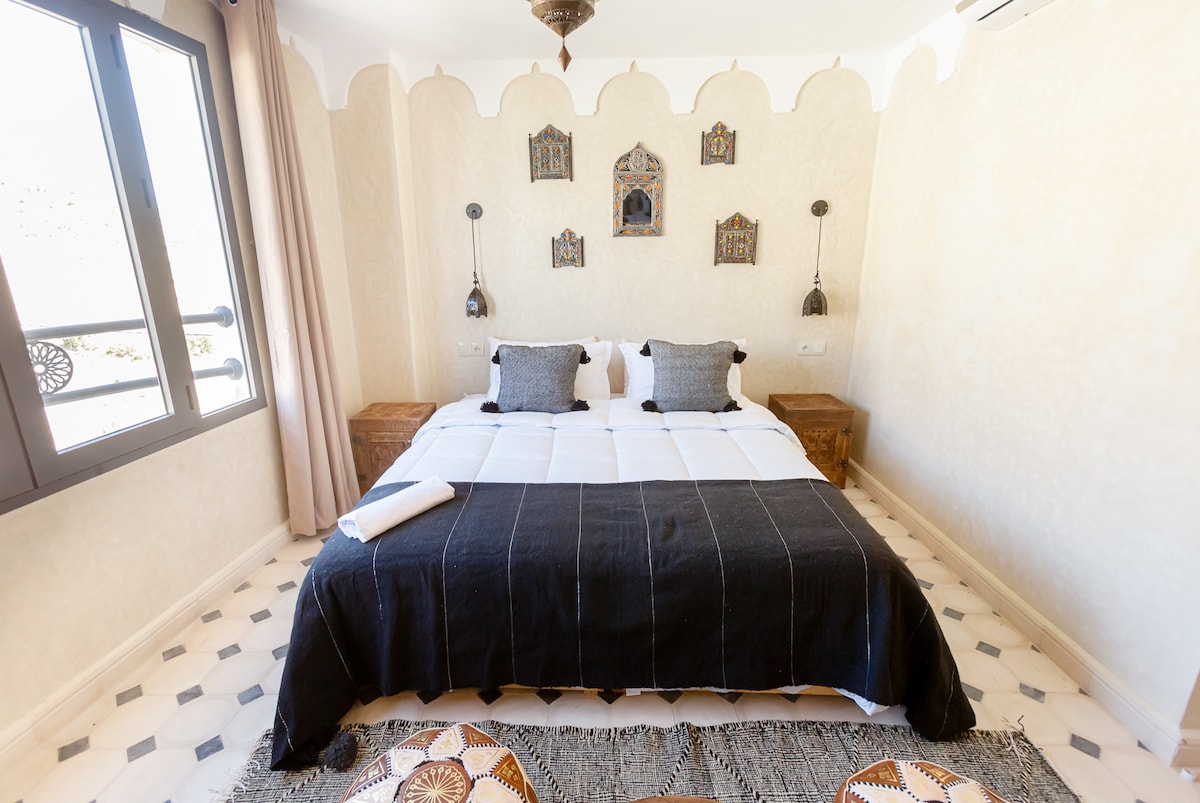 A spacious bedroom is featured, with a large bed adorned with black and white linens. Decorative wall hangings add character, while side tables are positioned on either side of the bed. Natural light filters through large windows with elegant drapes, illuminating the intricate tile flooring.