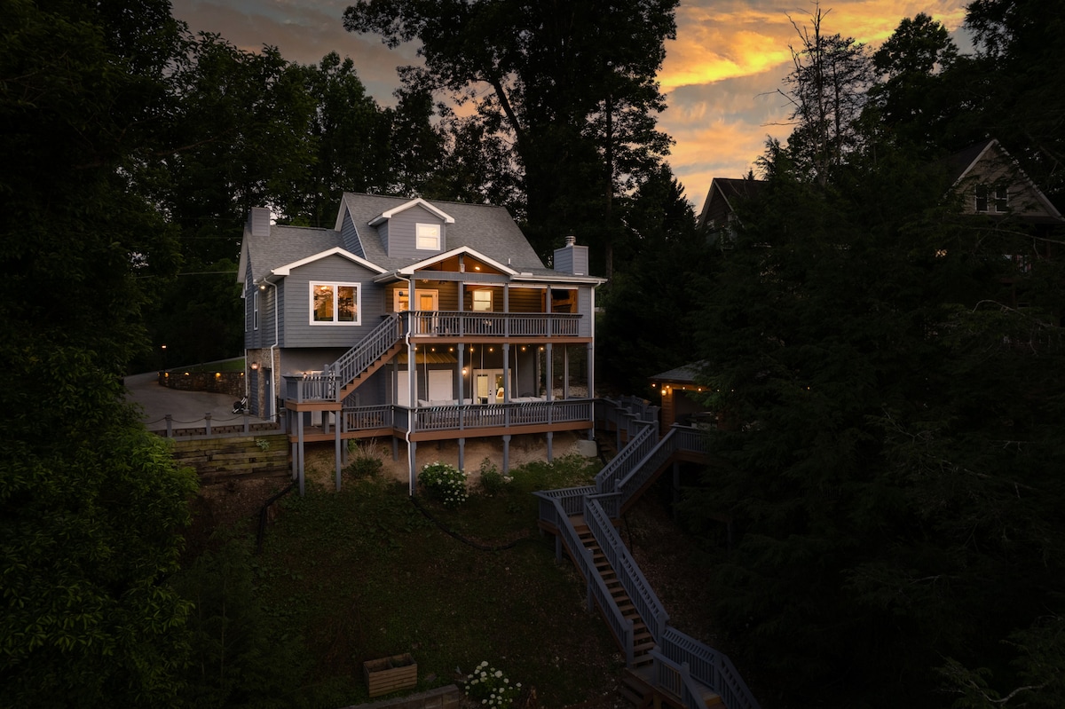 The exterior of a two-story lakefront home is captured during twilight, showcasing multiple decks and a spacious entrance. The house features large windows reflecting ambient light, surrounded by lush greenery and outdoor lighting creating a welcoming entrance. The sky is softly illuminated with a sunset glow.