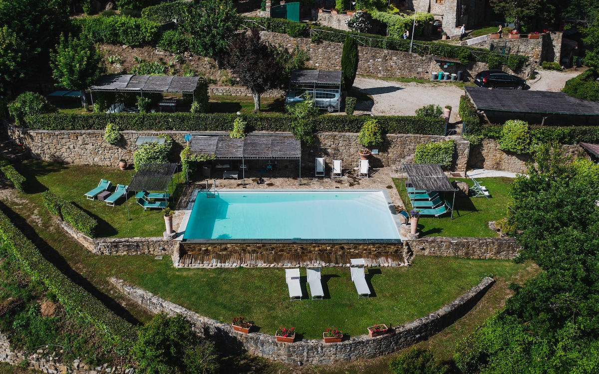 An outdoor pool area is surrounded by neatly trimmed hedges and stone walls. Sun loungers and shaded seating areas provide relaxation options. Lush greenery and flowering plants enhance the serene environment, creating an inviting space for guests to enjoy the tranquility.
