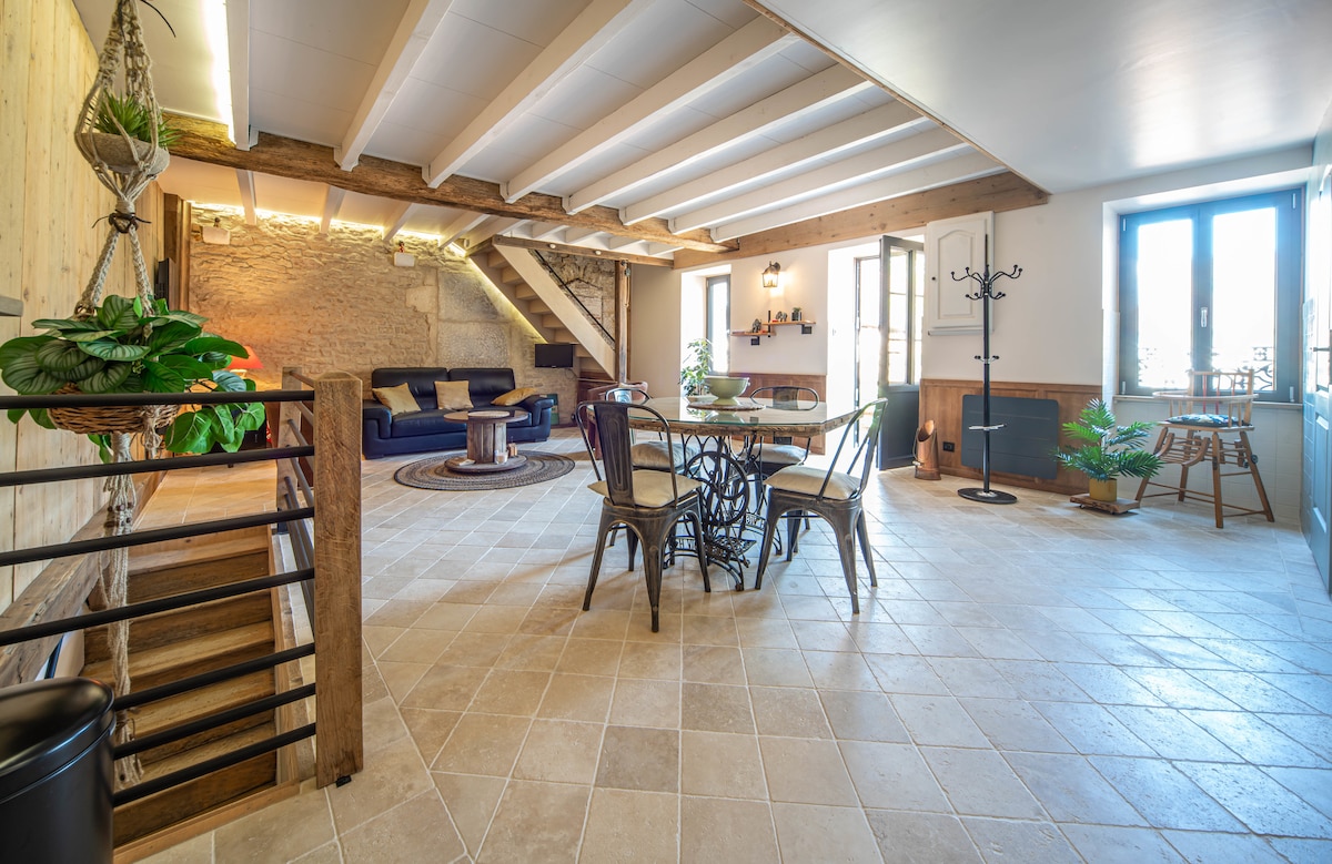 The main living area features exposed wooden beams and natural stone accents. A comfortable seating arrangement is paired with a round dining table surrounded by chairs. Sunlight enters through large windows, illuminating the tiled floor, and enhancing the spaciousness of the room.