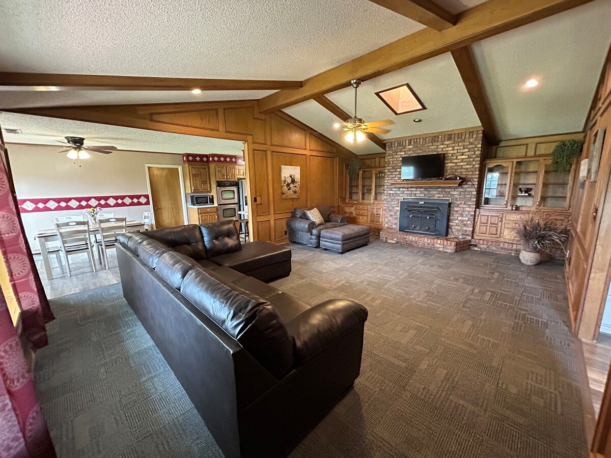 A spacious living area features wooden beams and a brick fireplace, creating a cozy focal point. Two dark sofas are arranged for relaxation, while a large window allows natural light to fill the space. An adjoining dining area is visible in the background.
