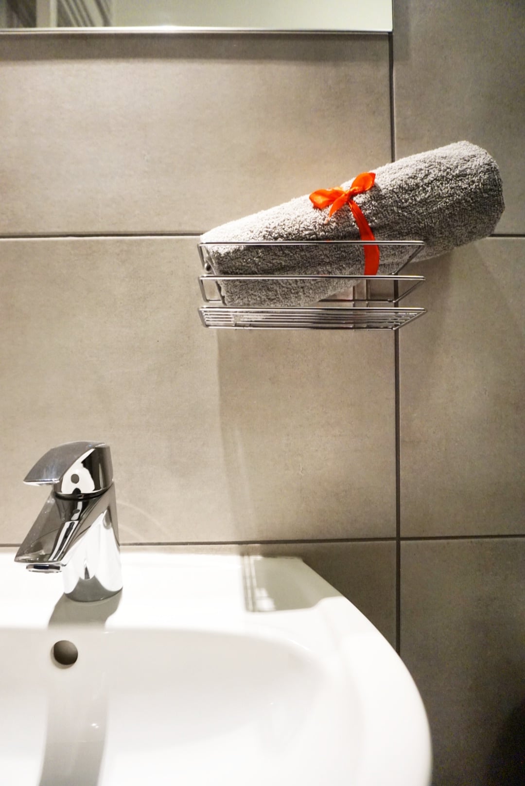 A modern bathroom sink is featured with a sleek faucet reflecting light. A neatly rolled grey towel tied with a red ribbon is placed on a metal shelf above the sink, complemented by textured grey wall tiles.