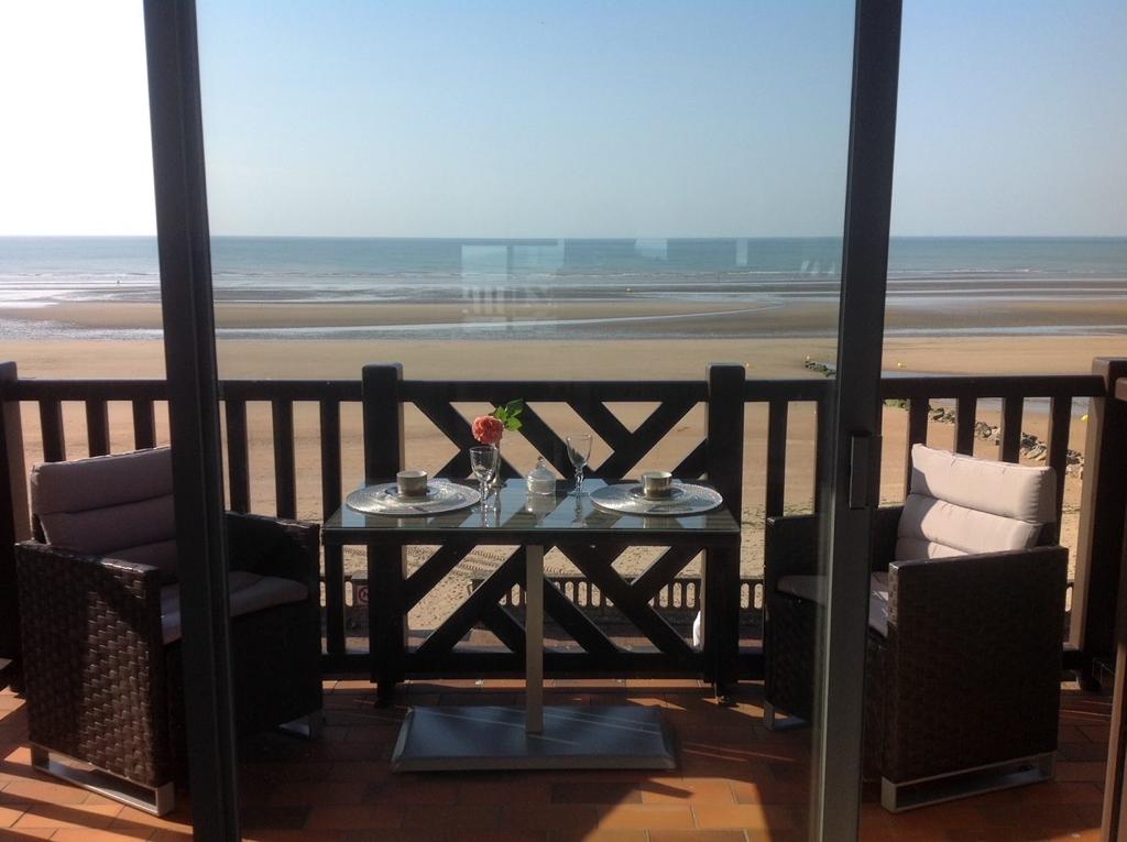 A balcony is presented with a table set for two, featuring elegant place settings and a flower centerpiece. Two armchairs provide comfortable seating, with a view of the expansive beach and sea in the distance. The glass doors enhance the connection to the outdoor space.