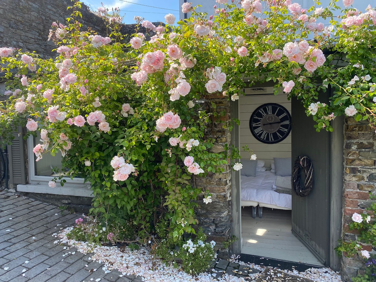 An inviting entrance is framed by blooming pink roses cascading over the doorway. Delicate petals cover the ground, leading to a light-filled interior featuring a cozy bed visible through the open door, creating a seamless transition between the garden and inside.