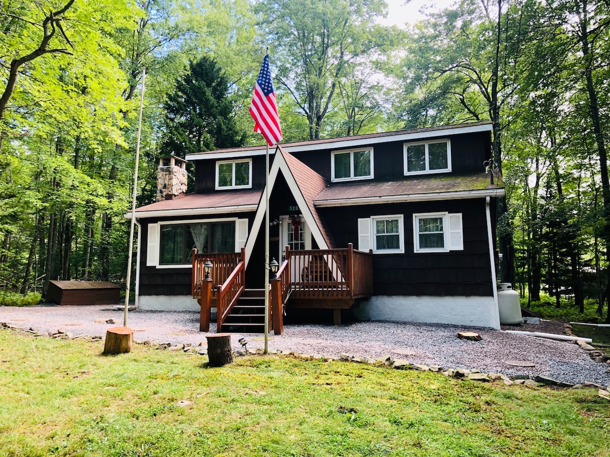 A two-story cabin is nestled among trees, featuring a wood exterior with white trim. A front deck with a railing is visible, leading to the entrance. An American flag waves proudly from a nearby pole, adding a touch of charm to the peaceful surroundings.