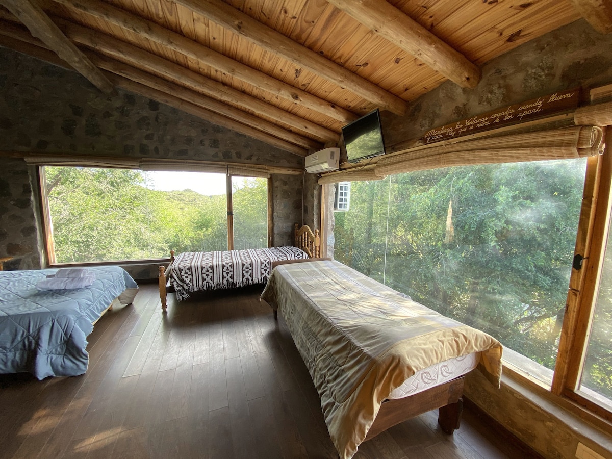 A spacious bedroom features wooden beams and stone walls, with two beds positioned near large windows that offer views of the surrounding greenery. A flat-screen television is mounted on the wall, and light-colored bedding complements the natural wood elements.