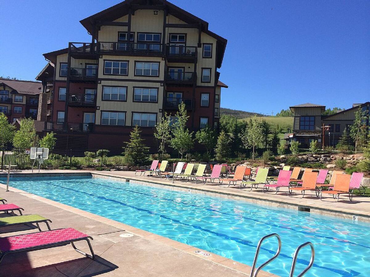 A large swimming pool is surrounded by brightly colored lounge chairs arranged on a patio. The clear blue water of the pool reflects the bright sky. Green trees are visible in the background, contrasting with the architecture of the nearby building.