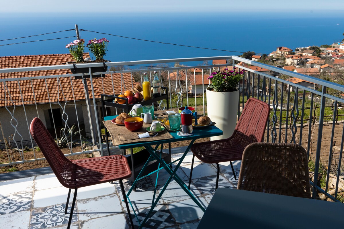 A terrace table is set for breakfast, featuring a variety of foods including fruits, pastries, and beverages. Two chairs are positioned around the table, with a backdrop of the sea visible in the distance. Flower pots add a touch of color to the setting.