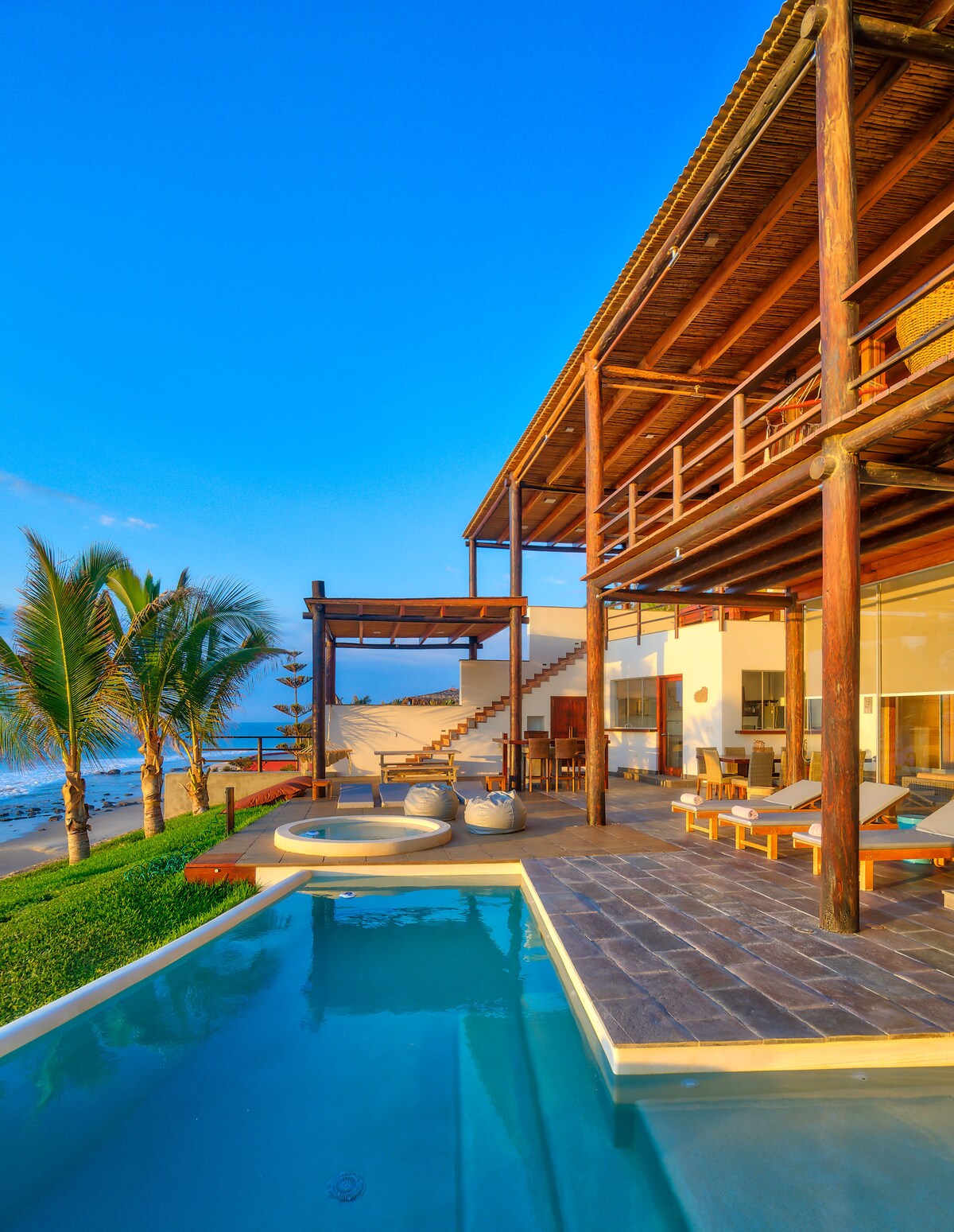A multi-level beachfront villa is depicted, showcasing a private pool with a clear blue surface. Outdoor seating areas with loungers are present near lush greenery, and large windows reflect the ocean views. Steps lead up to a covered terrace, enhancing the connection to the surroundings.