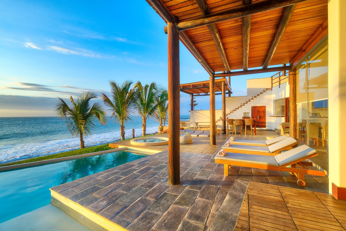 An outdoor terrace features sun loungers set near a serene pool, surrounded by palm trees and overlooking the ocean. A wooden dining area is positioned nearby, with steps leading to another level of the villa. Clear blue skies complement the tranquil seaside setting.