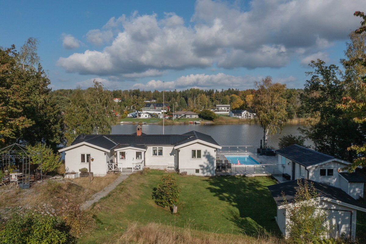 An elevated view of the villa is captured, showing a spacious lawn bordered by trees. The swimming pool glimmers nearby, with the lake reflecting the sky and surrounding landscapes. Charming residential houses dot the background, indicating a serene neighborhood setting.