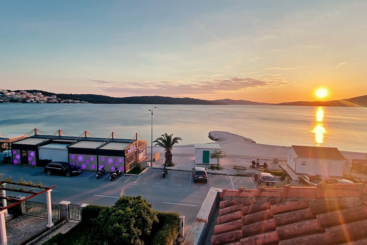 A scenic view captures the tranquil coastline at sunset. The sun is low on the horizon, casting warm tones across the water. Beachfront buildings and palm trees are visible, alongside parked vehicles and a sandy beach area.
