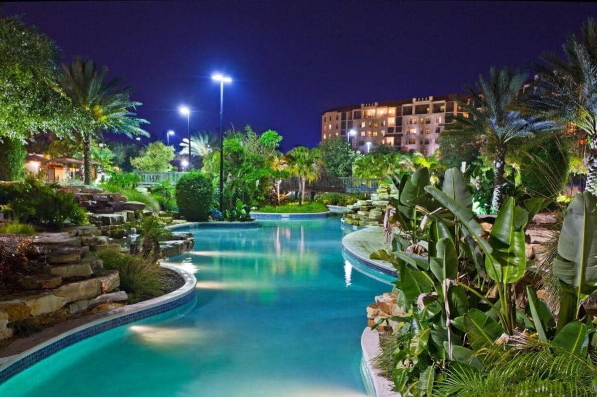 A winding lazy river flows through lush landscaping, surrounded by palm trees and vibrant greenery. Soft lighting illuminates the water, creating a tranquil atmosphere. In the background, a multi-story building stands amidst the resort's natural surroundings.