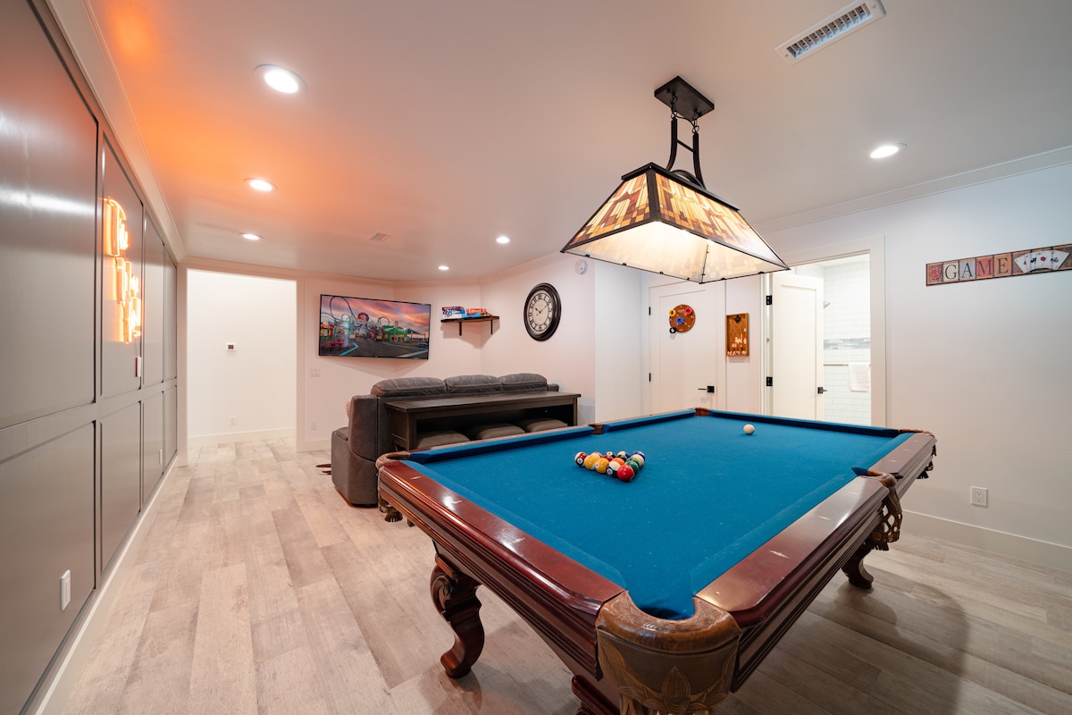 A game room features a classic pool table with colorful balls arranged on the felt. Soft lighting is provided by a decorative overhead fixture. A comfortable sofa is situated against the wall. Decorative artwork and a clock are displayed, enhancing the space's inviting ambiance.