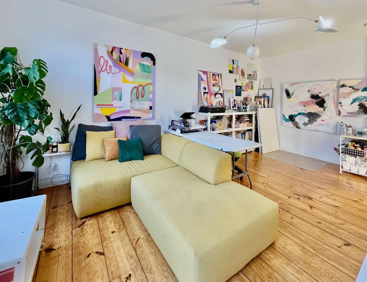 A spacious living area features a large yellow sectional sofa and a mix of vibrant wall art. Natural light floods the room, highlighting the original wooden floors. A potted plant adds a touch of greenery, while a worktable is positioned nearby, creating an inviting studio atmosphere.