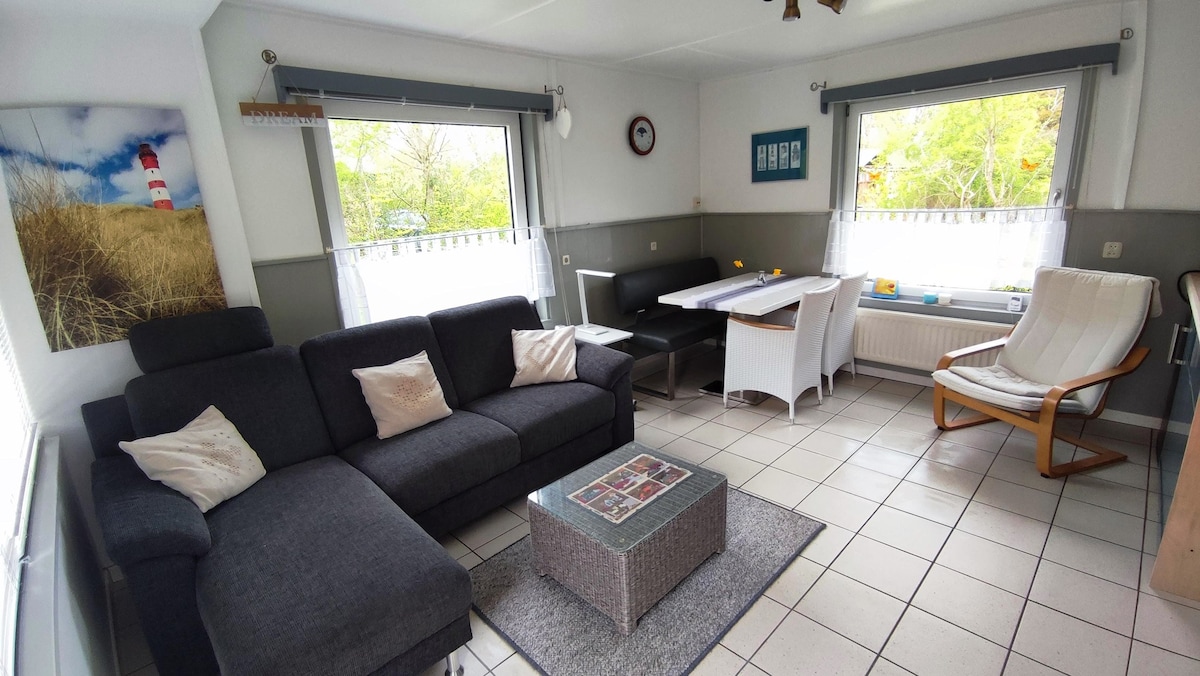 A comfortable living area is presented with a dark sectional sofa, accompanied by decorative pillows. A light wood coffee table is at the center, and a dining table with white chairs occupies one corner. Large windows allow natural light to fill the space.